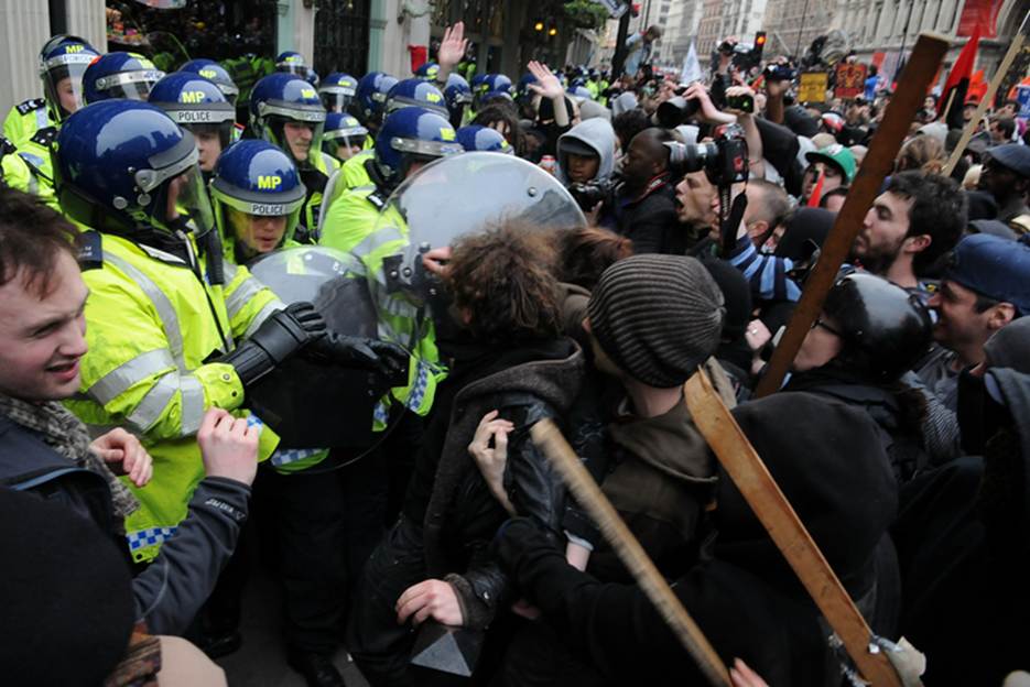 A crowded street scene with police in riot gear confronting protesters, some of whom are holding sticks. The police wear helmets with "MP" on them.